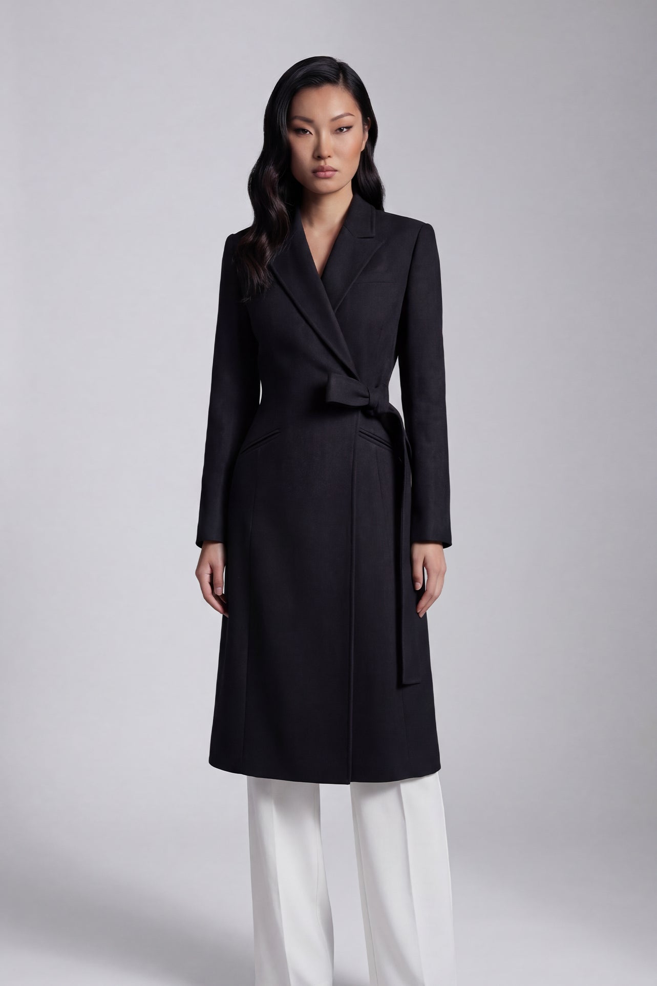 A professional woman wearing a sophisticated black long-sleeve coat dress, featuring an elegant tie-waist belt and notched lapels, styled over a white shirt for a polished executive business look.