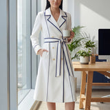 An elegant professional woman wearing a sophisticated white double-breasted coat dress featuring sharp navy blue contrast piping and a matching waist belt for a tailored executive look