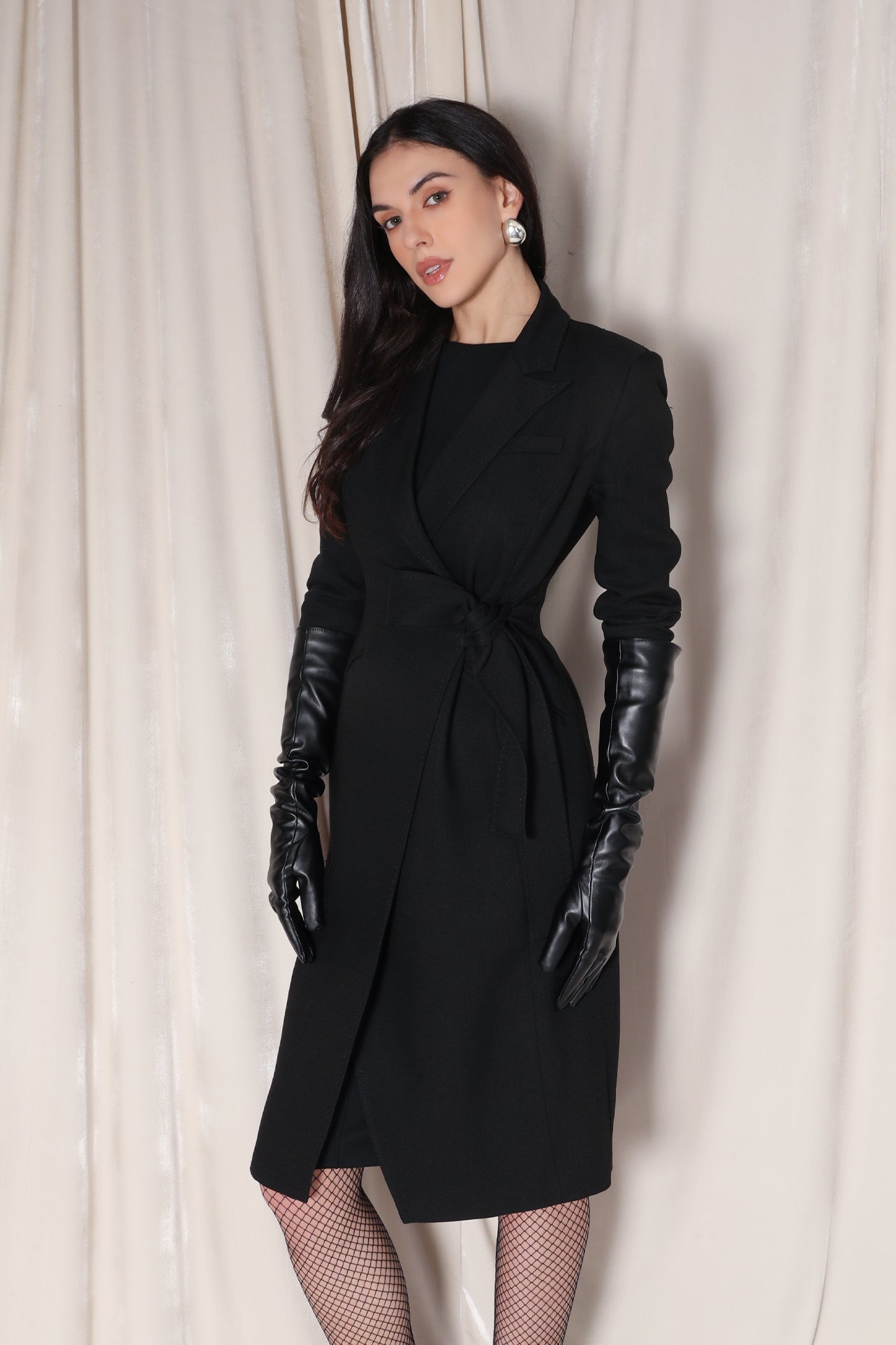 A professional woman wearing a sophisticated black long-sleeve coat dress, featuring an elegant tie-waist belt and notched lapels, styled over a white shirt for a polished executive business look.