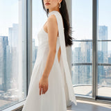 An elegant woman wearing a minimalist white spaghetti-strap A-line midi dress styled with a matching flowing neck scarf against a scenic Mediterranean blue sea and white architecture background