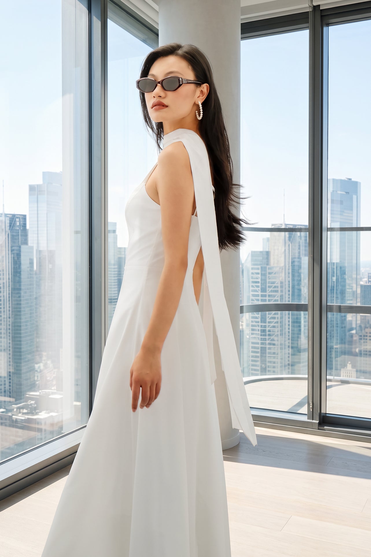 An elegant woman wearing a minimalist white spaghetti-strap A-line midi dress styled with a matching flowing neck scarf against a scenic Mediterranean blue sea and white architecture background