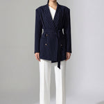 A professional woman wearing a sophisticated navy blue pinstripe blazer jacket, featuring a longline silhouette with a matching tie-waist belt and gold earring accents for an elegant executive office look.