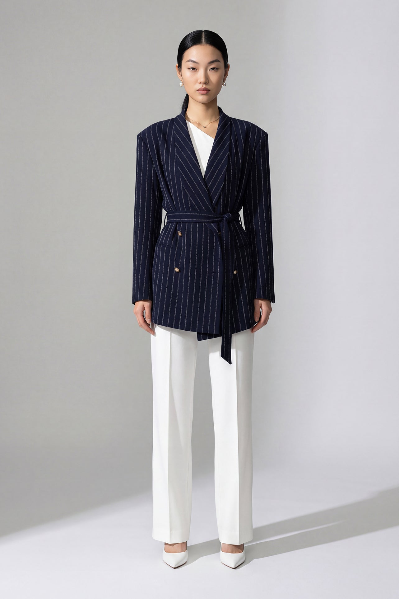 A professional woman wearing a sophisticated navy blue pinstripe blazer jacket, featuring a longline silhouette with a matching tie-waist belt and gold earring accents for an elegant executive office look.