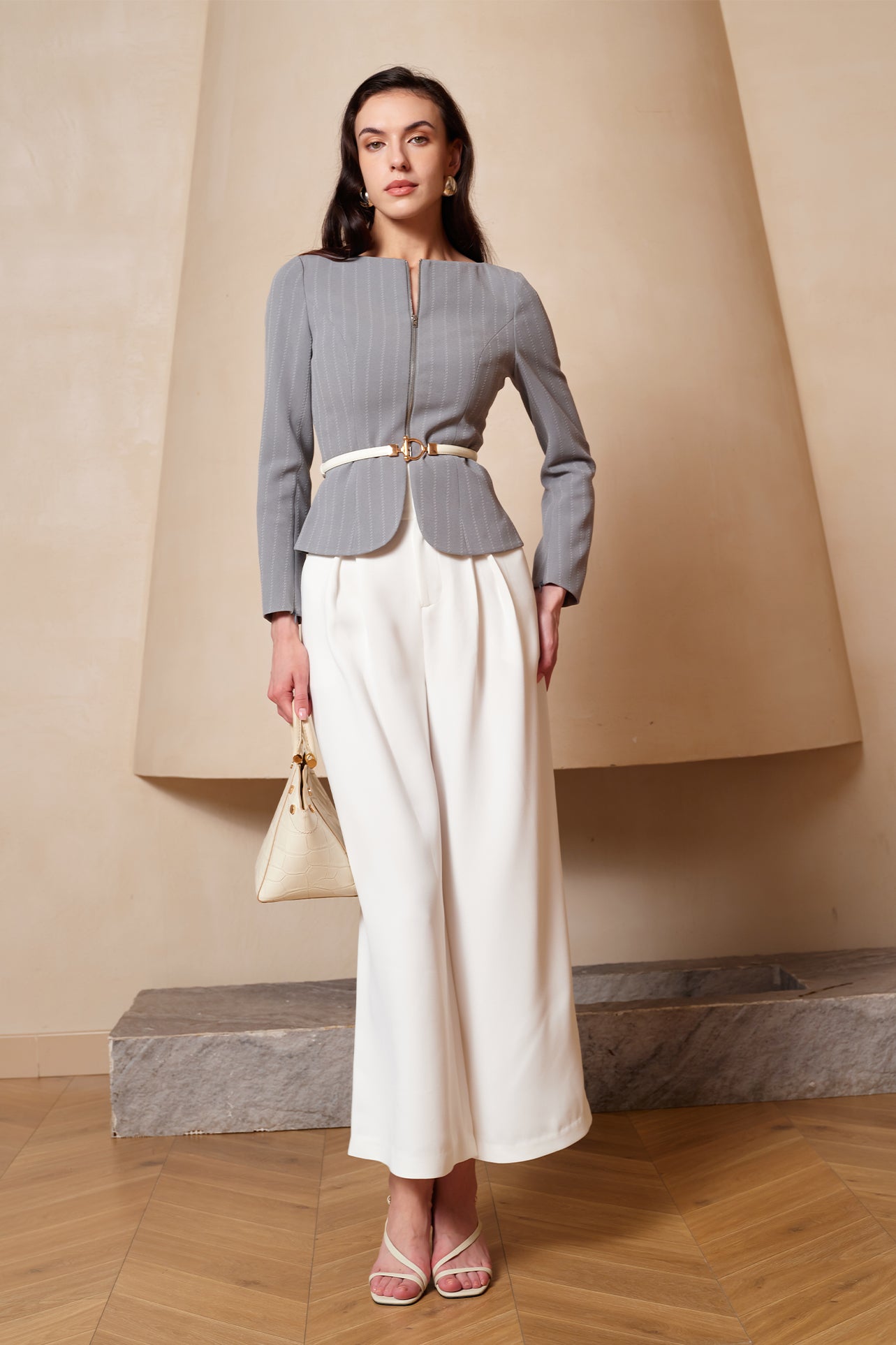 A professional woman wearing a sophisticated grey pinstripe zip-up blazer, featuring a thin white belt with a gold buckle and a tailored slim fit for a modern office look.