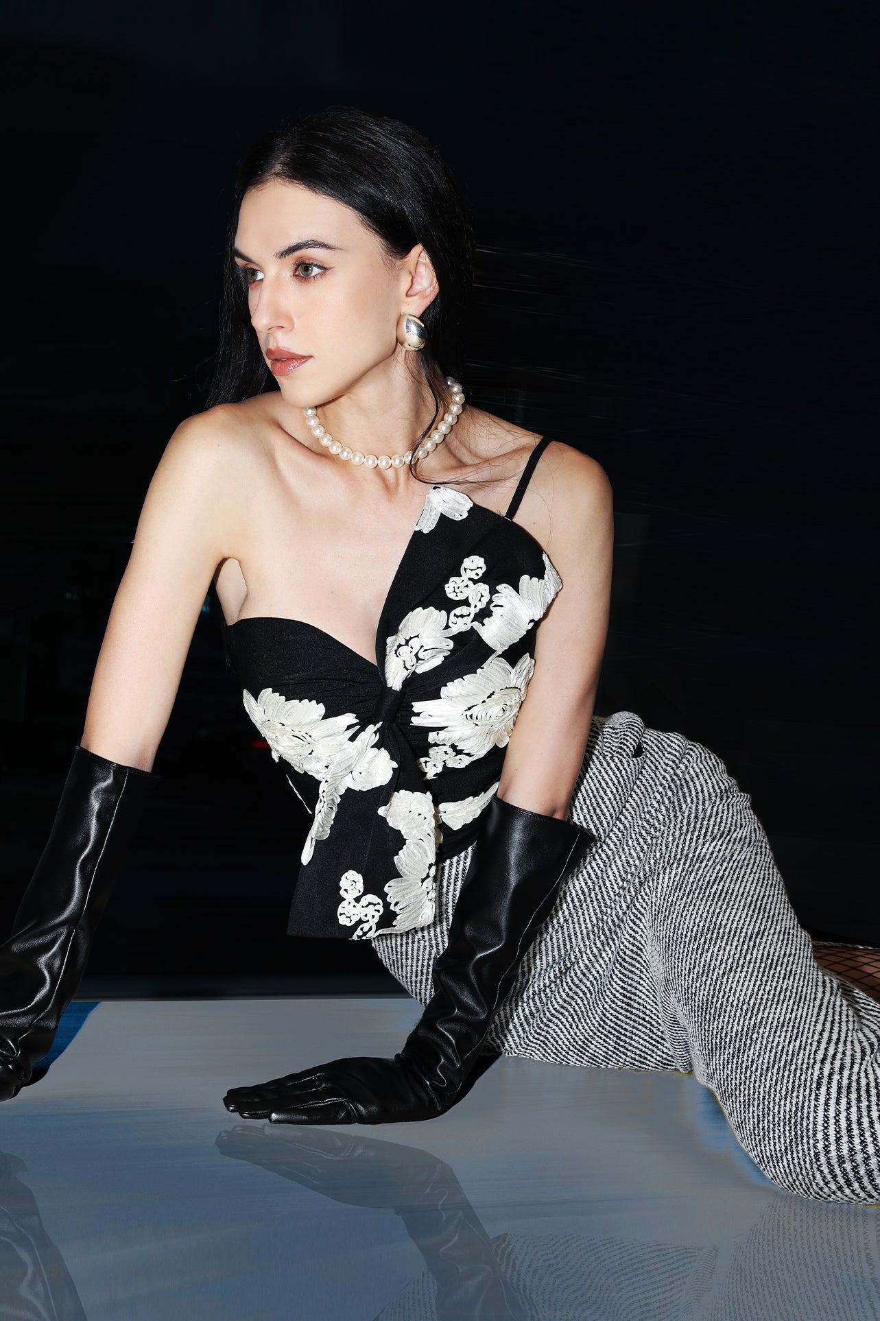 A professional woman wearing a sophisticated one-shoulder black and white floral print top, featuring a dramatic oversized bow detail on the shoulder, paired with white high-waisted trousers for an elegant evening look.