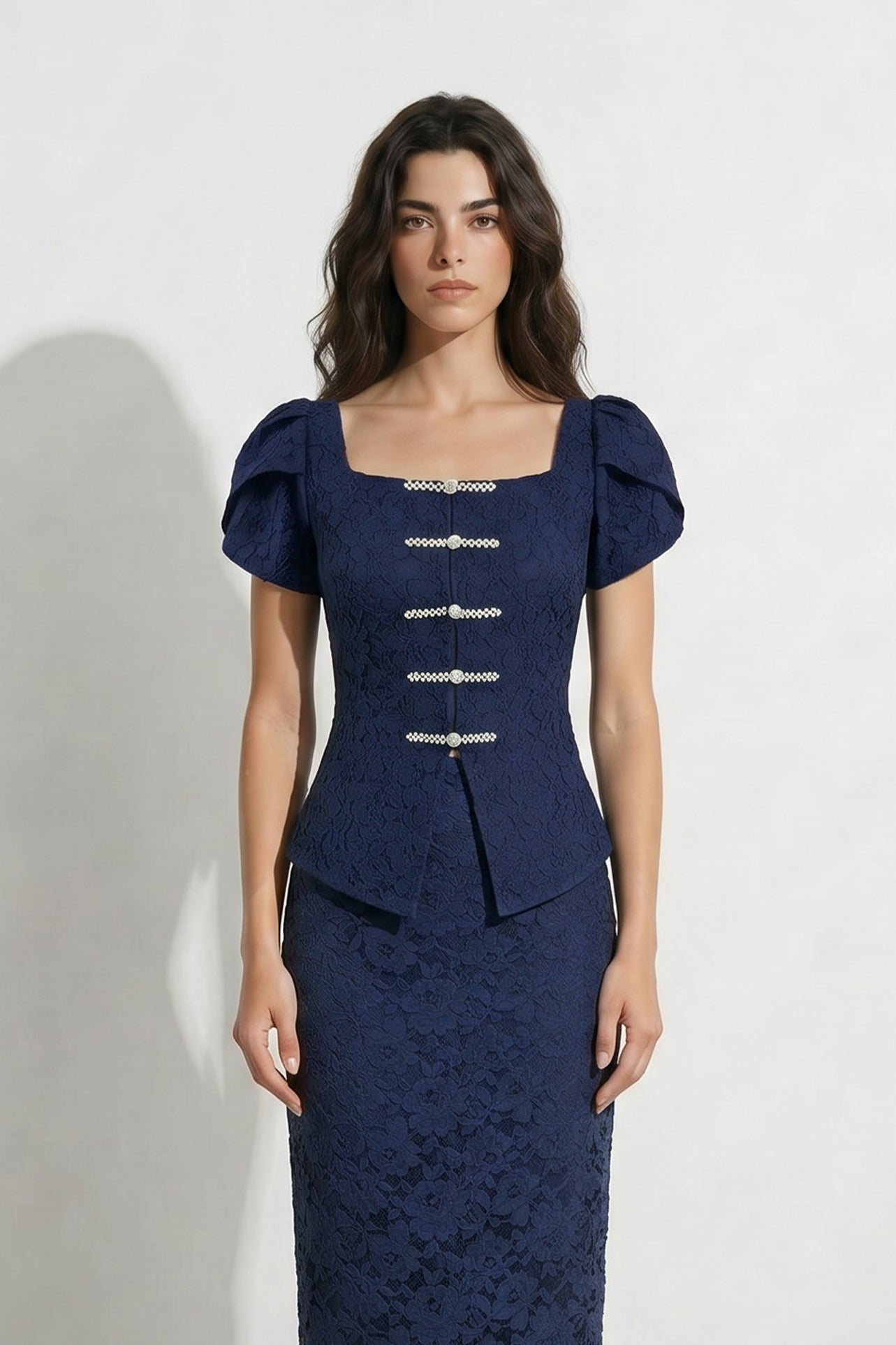 A professional woman modeling an elegant navy blue lace short top, featuring a retro square neckline, romantic short puff sleeves, and a series of pearl and rhinestone frog closures on the front with a tailored triangular split hem, paired with light blue high-waisted jeans.