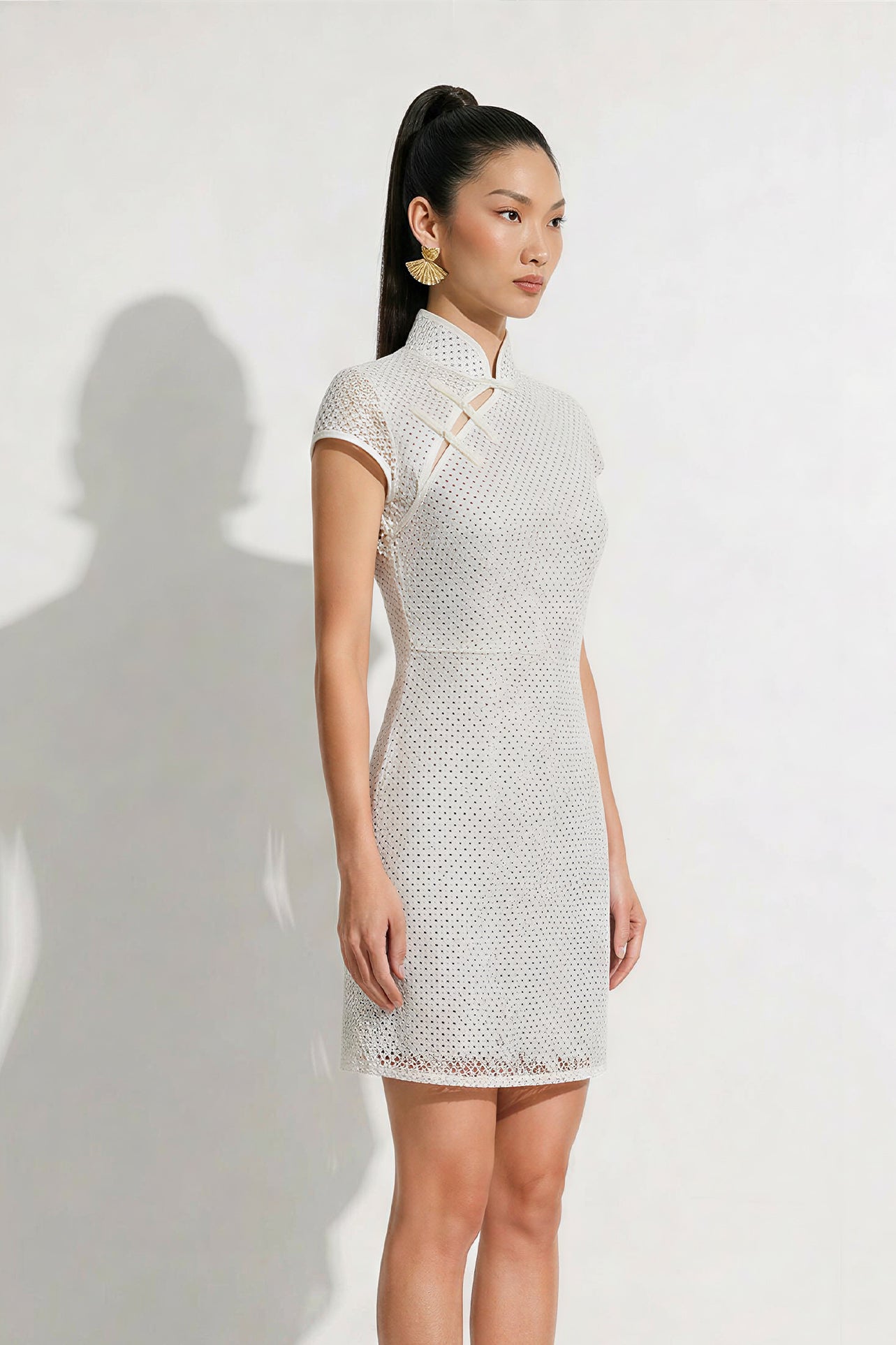 A professional woman modeling an elegant white lace modern qipao dress, featuring an intricate eyelet textured fabric, a sophisticated mandarin collar with a traditional knot button, and slimming cap sleeves.