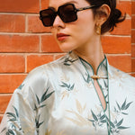 A professional woman modeling an elegant light sage green silk wrap jacket, featuring dark green bamboo leaf prints, a sophisticated mandarin collar with keyhole detail, and wide kimono sleeves.
