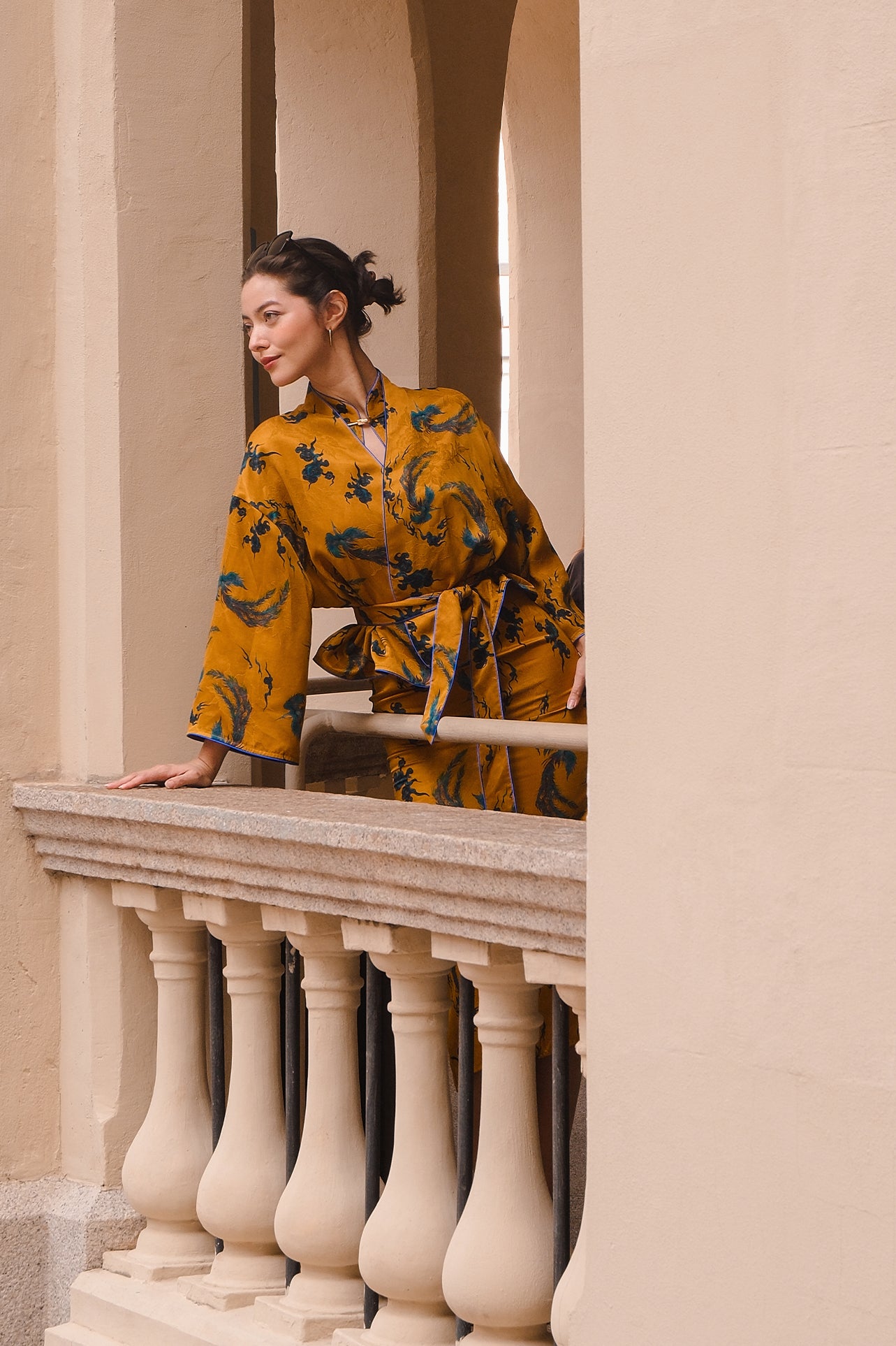 A beautiful woman modeling an elegant mustard yellow modern Chinese jacket, featuring intricate blue phoenix embroidery, a sophisticated mandarin collar, and wide kimono sleeves with a matching waist tie.