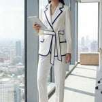 An elegant professional woman wearing a tailored white blazer with navy blue contrast piping, featuring a self-tie belt to define the waist for a smart-casual office look