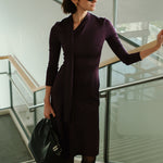 An elegant woman wearing a sophisticated deep plum-purple long-sleeve knit midi dress featuring a draped high-neck tie scarf and a dramatic asymmetrical flounce mermaid hemline.