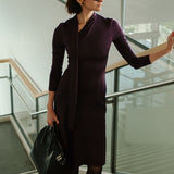 An elegant woman wearing a sophisticated deep plum-purple long-sleeve knit midi dress featuring a draped high-neck tie scarf and a dramatic asymmetrical flounce mermaid hemline.