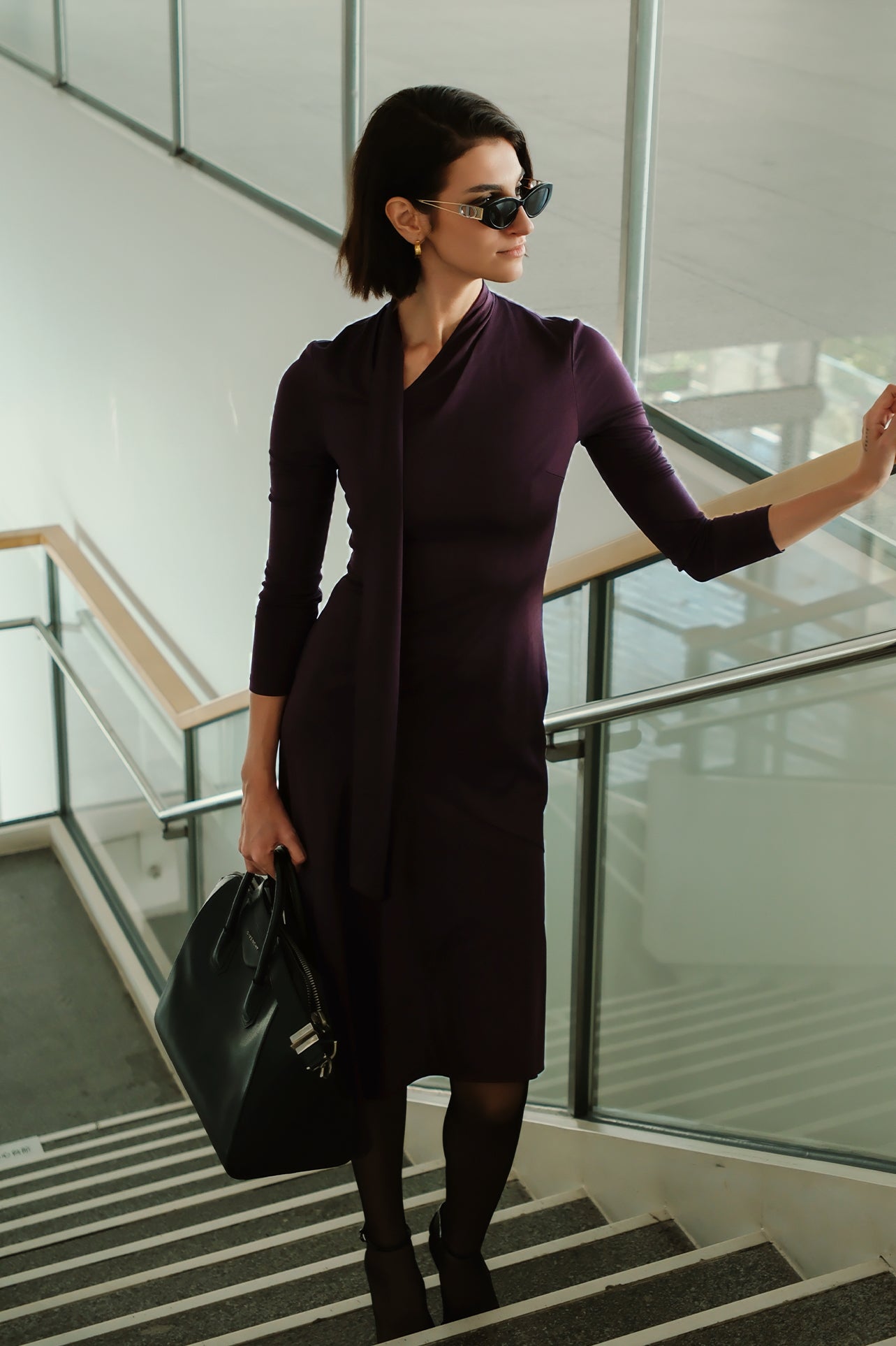 An elegant woman wearing a sophisticated deep plum-purple long-sleeve knit midi dress featuring a draped high-neck tie scarf and a dramatic asymmetrical flounce mermaid hemline.