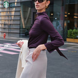 A chic woman wearing an elegant deep plum long-sleeve blouse with an asymmetric high-neck scarf tie, neatly tucked into a high-waisted black tweed skirt embellished with subtle sparkling sequins.