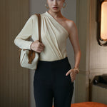A stylish Asian woman modeling a sophisticated cream-colored asymmetric one-shoulder top featuring a single long sleeve, elegant diagonal draped pleats, and styled with high-waisted black trousers.