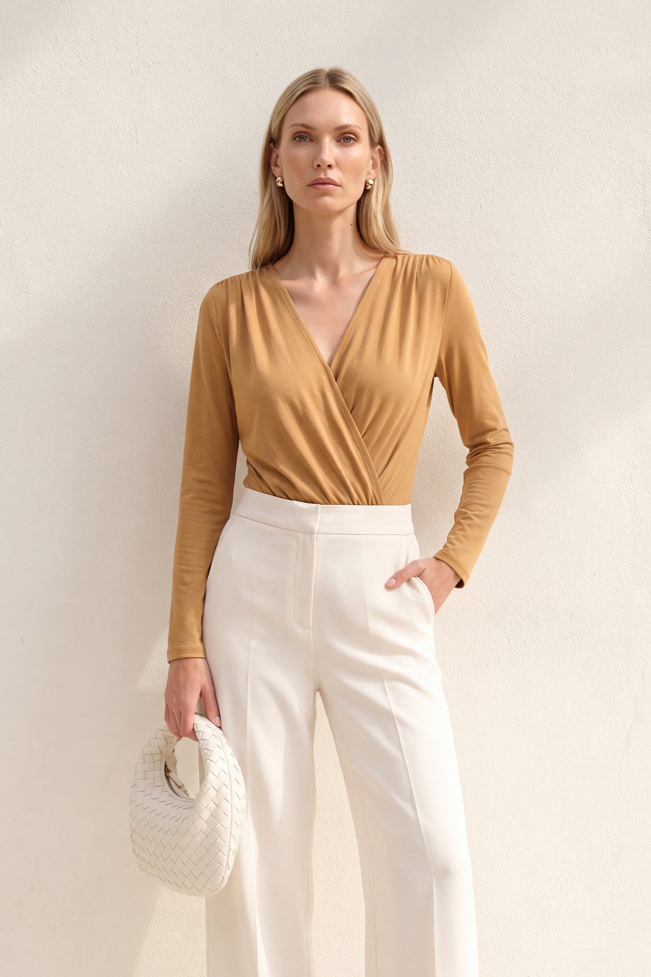 A style comparison of two elegant long-sleeve V-neck wrap tops featuring delicate shoulder pleating and a sleek tucked-in fit; the left in warm camel paired with cream trousers, and the right in deep burgundy paired with black trousers.