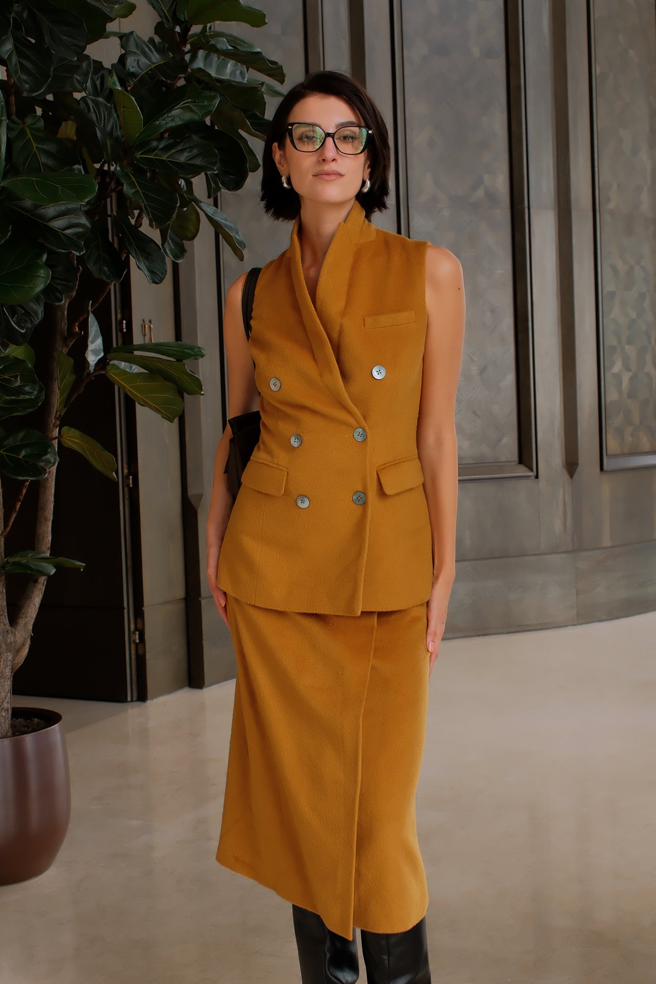 A chic professional woman wearing a tailored camel double-breasted longline suit vest featuring classic notch lapels and flap pockets, layered over a soft beige turtleneck sweater for a sophisticated quiet luxury look.
