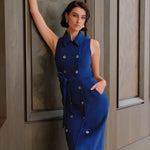 An elegant woman wearing a sophisticated royal cobalt blue trench dress featuring a structured turn-down collar, a cropped capelet overlay, a double-breasted button front, and a cinched self-tie belted waist.