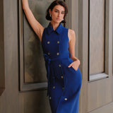 An elegant woman wearing a sophisticated royal cobalt blue trench dress featuring a structured turn-down collar, a cropped capelet overlay, a double-breasted button front, and a cinched self-tie belted waist.