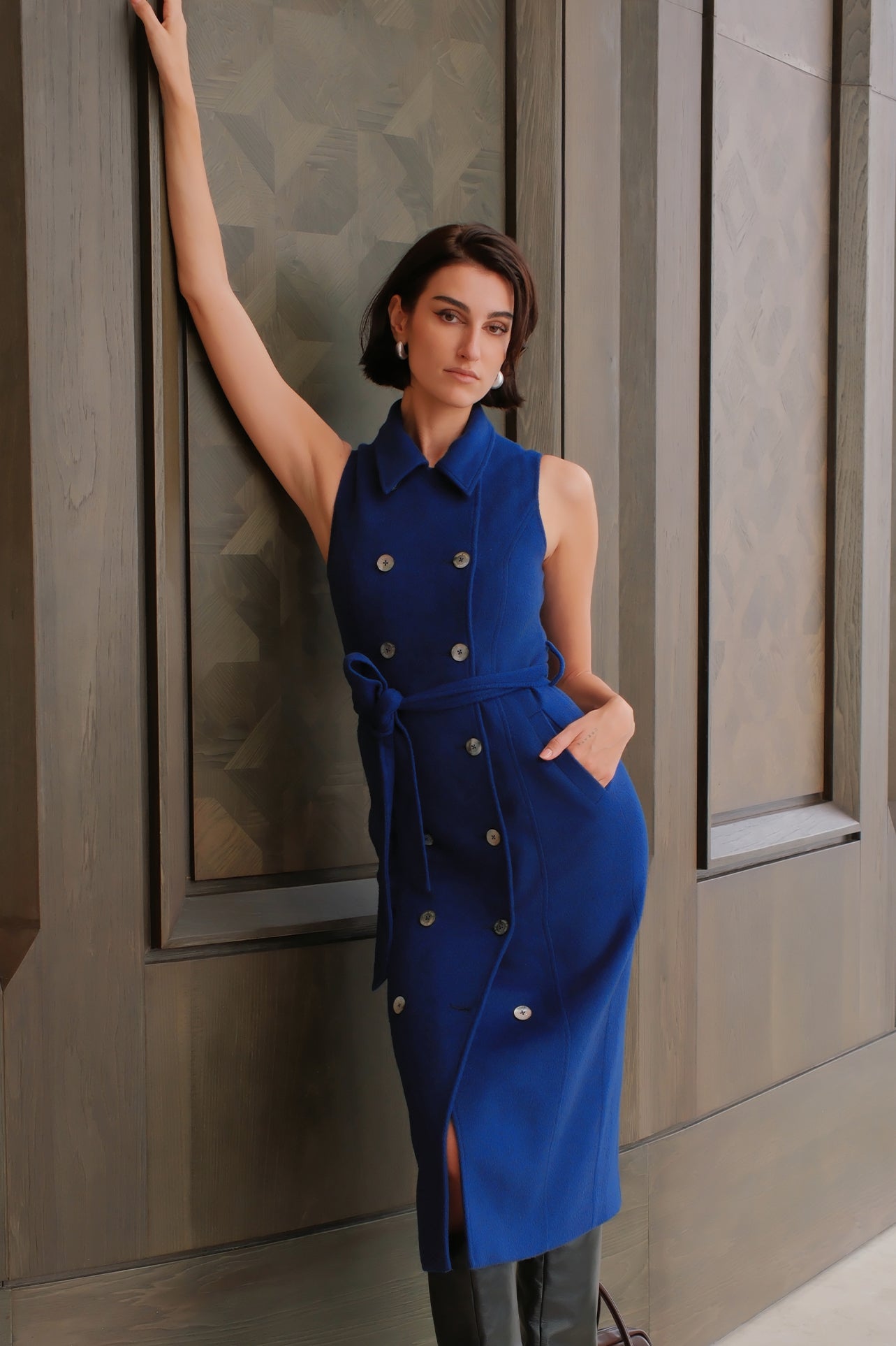 An elegant woman wearing a sophisticated royal cobalt blue trench dress featuring a structured turn-down collar, a cropped capelet overlay, a double-breasted button front, and a cinched self-tie belted waist.