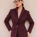 A professional woman wearing glasses and an elegant deep burgundy skirt suit featuring a sculptural stand-up lapel blazer with a single-button closure, tailored flap pockets, and a matching high-waisted pencil skirt.