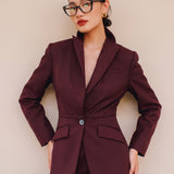 A professional woman wearing glasses and an elegant deep burgundy skirt suit featuring a sculptural stand-up lapel blazer with a single-button closure, tailored flap pockets, and a matching high-waisted pencil skirt.