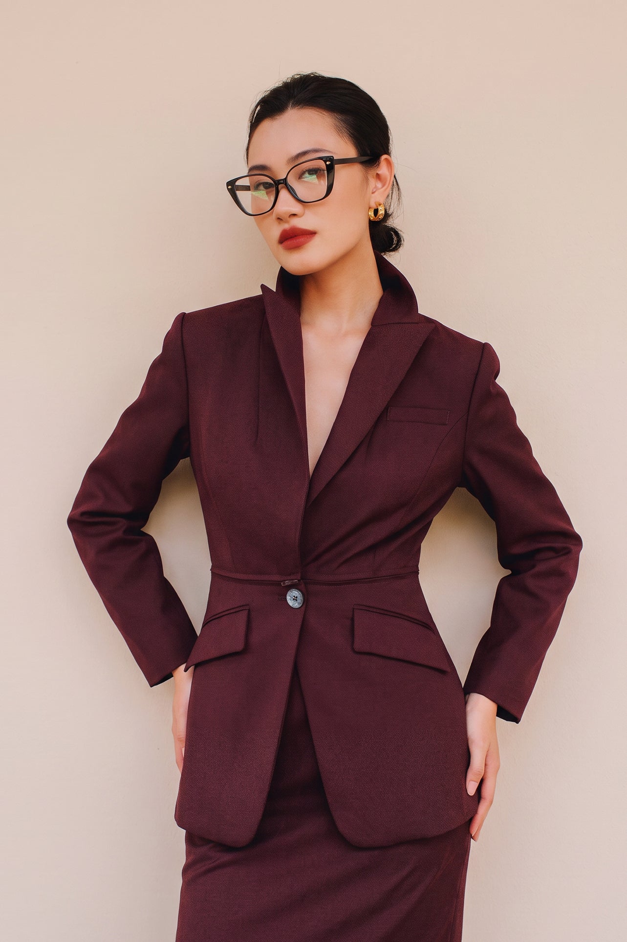 A professional woman wearing glasses and an elegant deep burgundy skirt suit featuring a sculptural stand-up lapel blazer with a single-button closure, tailored flap pockets, and a matching high-waisted pencil skirt.