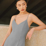 A stylish woman walking on a city street wearing an elegant light grey sheath midi dress with a sophisticated capelet sleeve design, round neckline, and side slit, carrying a black leather handbag.