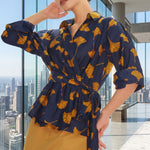 A stylish woman posing against a city skyline wearing a navy blue wrap blouse with golden ginkgo leaf print tied at the waist, paired with tailored mustard yellow wide-leg trousers.