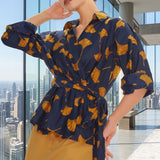A stylish woman posing against a city skyline wearing a navy blue wrap blouse with golden ginkgo leaf print tied at the waist, paired with tailored mustard yellow wide-leg trousers.