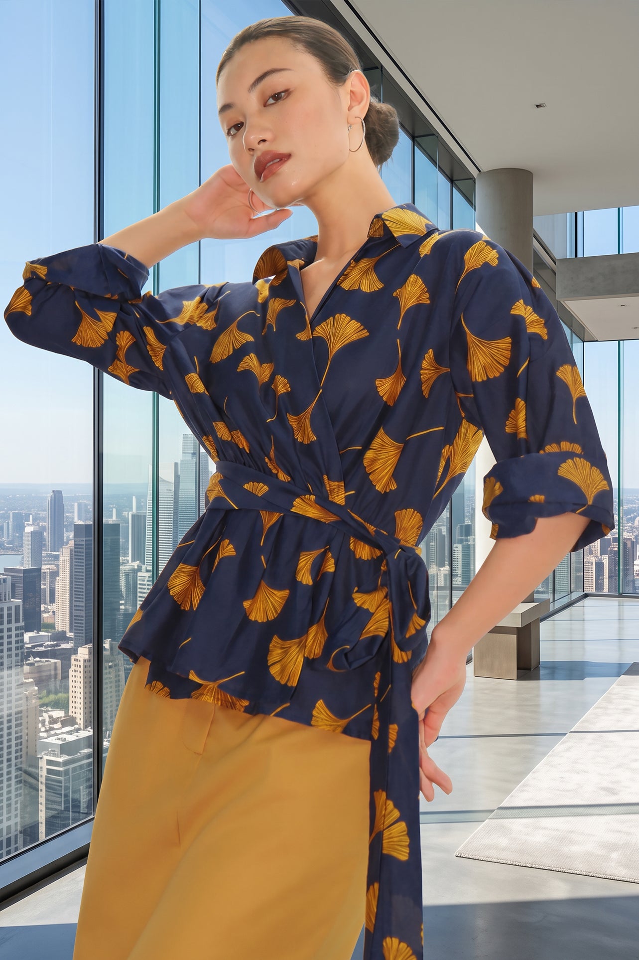 A stylish woman posing against a city skyline wearing a navy blue wrap blouse with golden ginkgo leaf print tied at the waist, paired with tailored mustard yellow wide-leg trousers.