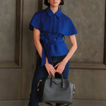A style comparison of two professional women wearing modern cobalt blue outerwear featuring self-tie waist belts; on the left, a tailored cropped capelet jacket paired with jeans, and on the right, a double-breasted sleeveless vest with white contrast buttons styled with black trousers.