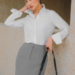 A stylish woman wearing a sophisticated grey pinstripe high-waisted midi skirt with a front slit, paired with a white button-up shirt rolled at the sleeves and black ankle boots for a minimalist corporate chic look.