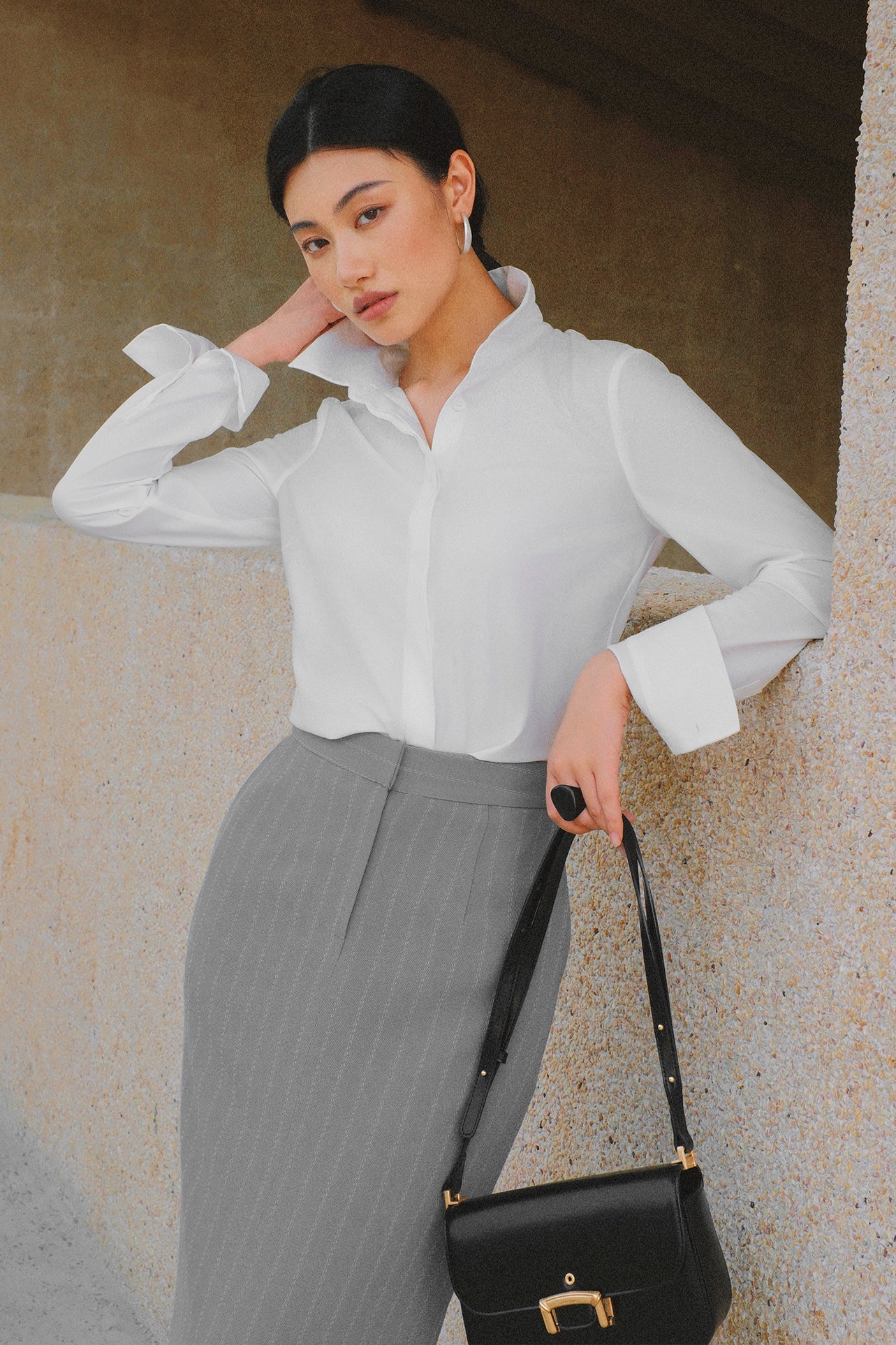 A stylish woman wearing a sophisticated grey pinstripe high-waisted midi skirt with a front slit, paired with a white button-up shirt rolled at the sleeves and black ankle boots for a minimalist corporate chic look.