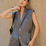 A style comparison showing two ways to wear a longline sleeveless blazer vest: on the left, a slate blue vest layered over a white shirt and cinched with a brown belt worn as a dress with knee-high boots; on the right, an oatmeal beige vest styled with flowing white wide-leg trousers.