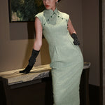 A stylish woman wearing sunglasses and holding a black folding fan, dressed in an elegant mint green sleeveless modern qipao dress featuring a mandarin collar with delicate button trim along the bodice and a tailored midi sheath silhouette.
