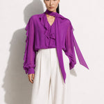 A style comparison of two professional corporate outfits featuring long-sleeve ruffled chiffon blouses with versatile tie-neck scarves; the left styled in purple with white wide-leg trousers, and the right in grey paired with a mustard yellow high-waisted pencil skirt.