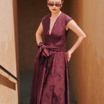 A chic woman wearing sunglasses and an elegant deep burgundy sleeveless V-neck A-line midi dress featuring textured jacquard fabric, delicate lace hemline, and a matching flowing neck scarf.