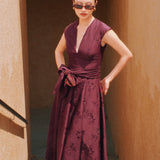 A chic woman wearing sunglasses and an elegant deep burgundy sleeveless V-neck A-line midi dress featuring textured jacquard fabric, delicate lace hemline, and a matching flowing neck scarf.