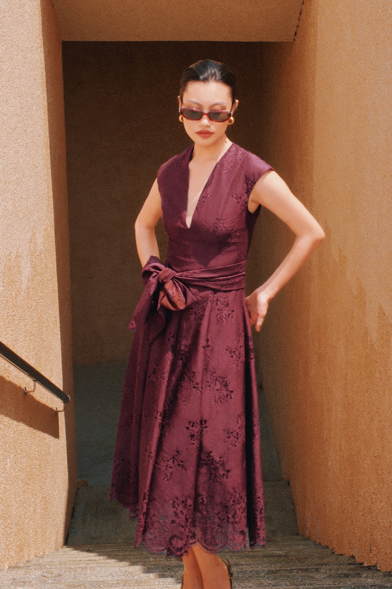 A chic woman wearing sunglasses and an elegant deep burgundy sleeveless V-neck A-line midi dress featuring textured jacquard fabric, delicate lace hemline, and a matching flowing neck scarf.