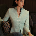 An elegant woman sitting on a modern chair wearing a sophisticated mint green textured suit featuring a cropped jacket with a mandarin collar, three white frog closures, and a matching high-waisted front-slit pencil skirt.