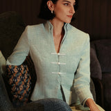 An elegant woman sitting on a modern chair wearing a sophisticated mint green textured suit featuring a cropped jacket with a mandarin collar, three white frog closures, and a matching high-waisted front-slit pencil skirt.