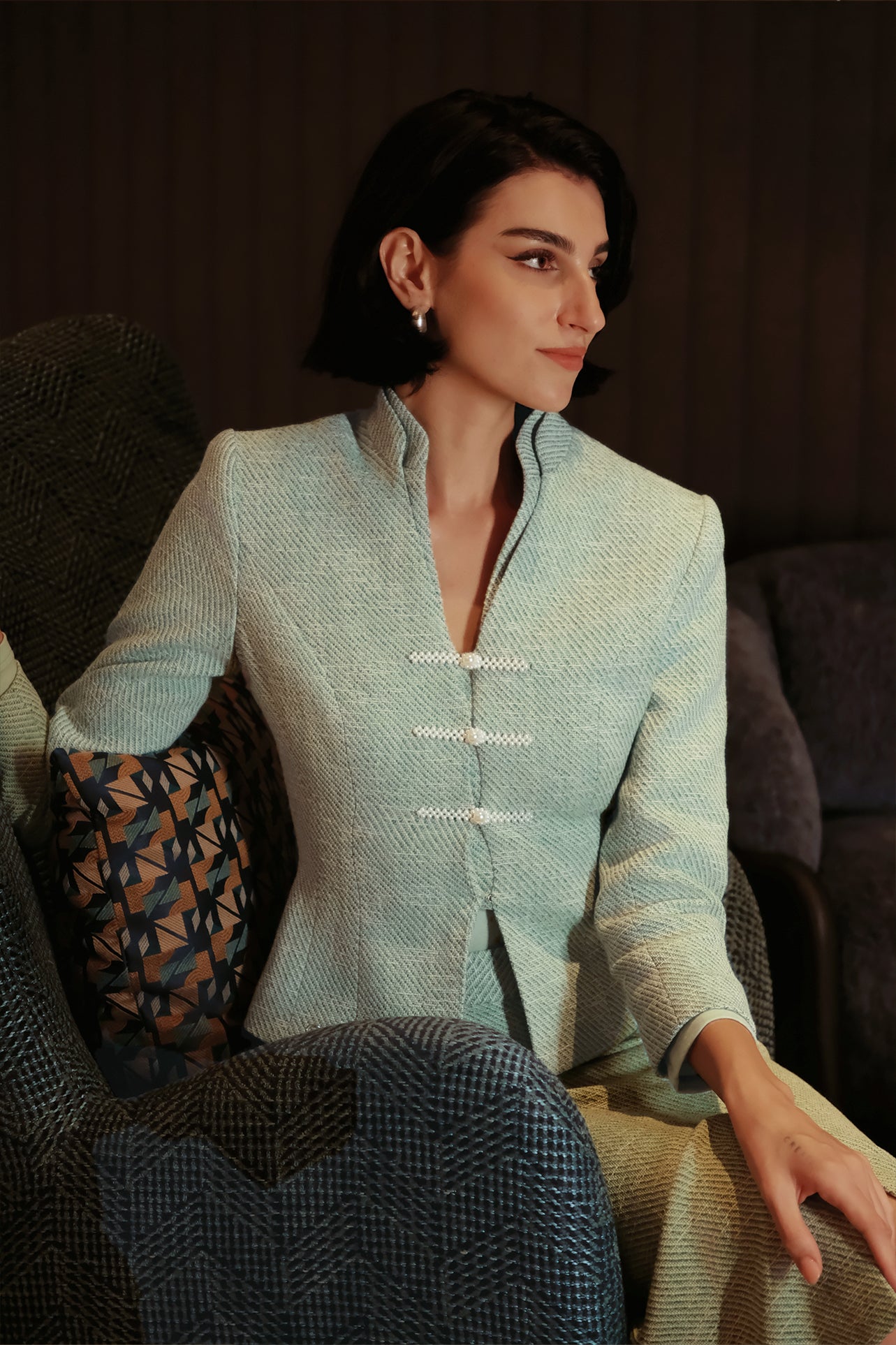 An elegant woman sitting on a modern chair wearing a sophisticated mint green textured suit featuring a cropped jacket with a mandarin collar, three white frog closures, and a matching high-waisted front-slit pencil skirt.