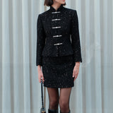 A detailed shot of a woman modeling a sophisticated black woven tweed mini skirt adorned with subtle shimmering crystals, tailored in a sleek high-waisted pencil cut.