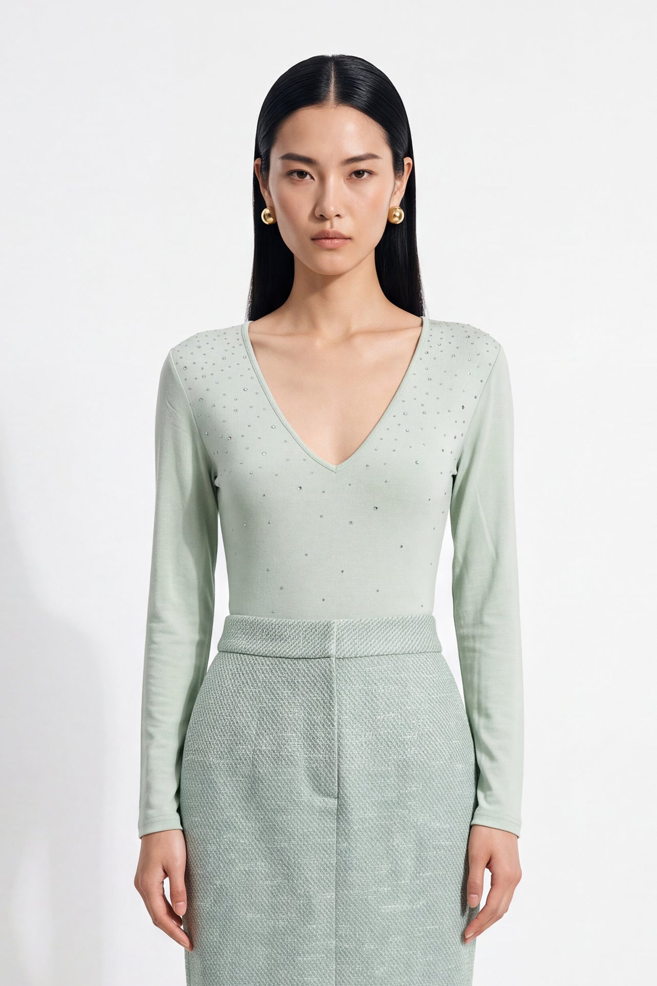 A style comparison of two elegant women's long-sleeve deep V-neck tops featuring subtle rhinestone crystal embellishments; shown on the left in nude camel tucked into tailored trousers, and on the right in pale mint green styled with a white skirt.