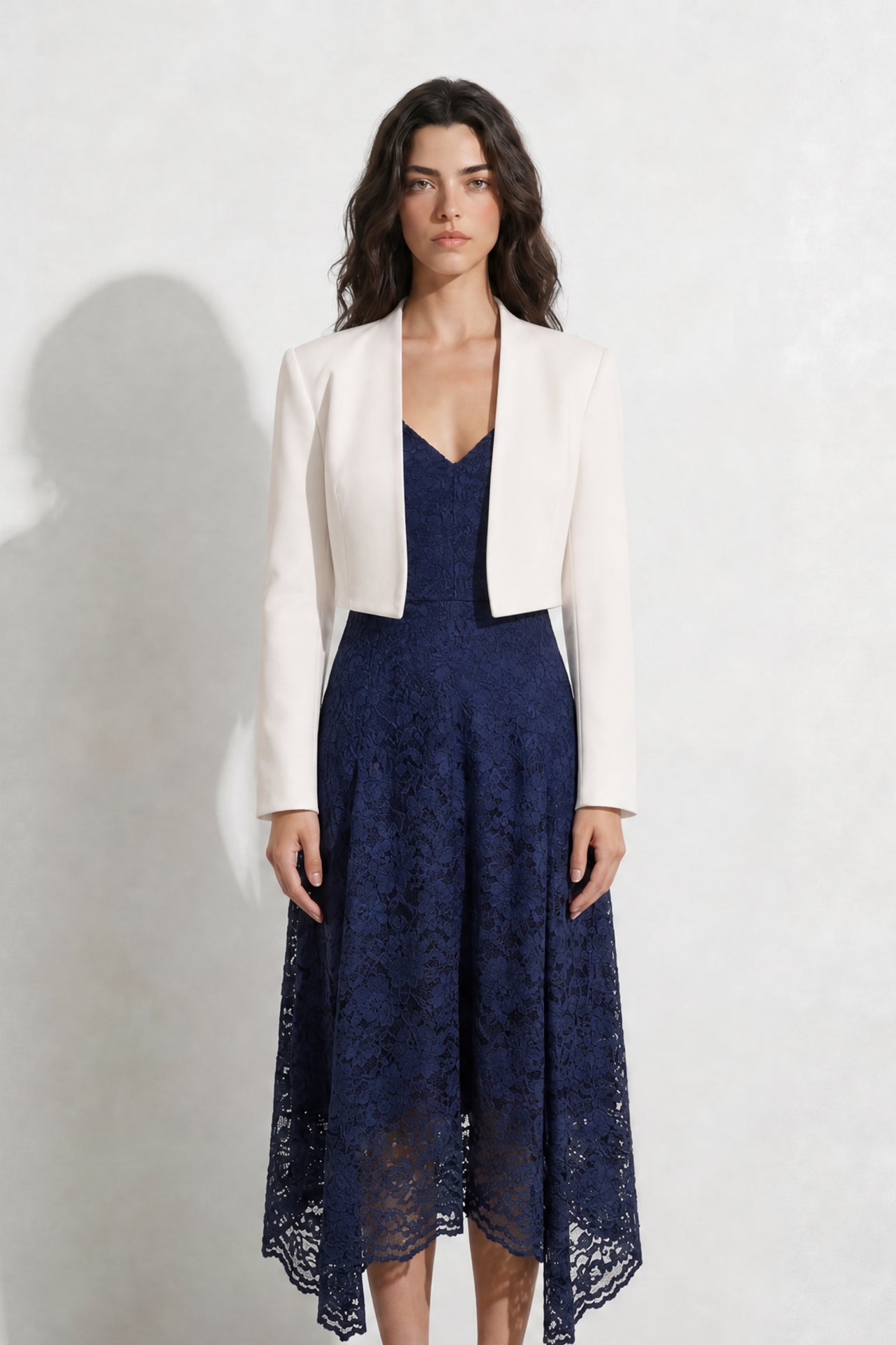 A professional woman modeling an elegant navy blue lace midi dress, featuring delicate V-neck spaghetti straps, a tailored fit-and-flare silhouette, and a beautiful flowing asymmetric handkerchief lace trim hem.