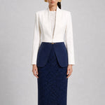 A professional model showcasing an elegant two-tone women's pantsuit, featuring a tailored cropped white blazer with a sharp deep V-neckline, paired with high-waisted navy blue trousers that have a unique curved overlay panel design and a golden metal hardware accent at the waist.