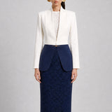 A professional model showcasing an elegant two-tone women's pantsuit, featuring a tailored cropped white blazer with a sharp deep V-neckline, paired with high-waisted navy blue trousers that have a unique curved overlay panel design and a golden metal hardware accent at the waist.