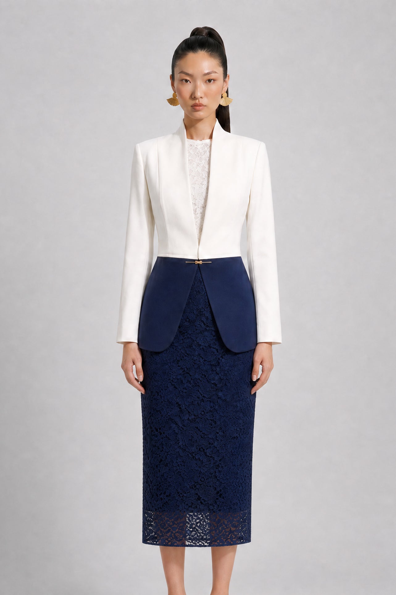 A professional model showcasing an elegant two-tone women's pantsuit, featuring a tailored cropped white blazer with a sharp deep V-neckline, paired with high-waisted navy blue trousers that have a unique curved overlay panel design and a golden metal hardware accent at the waist.