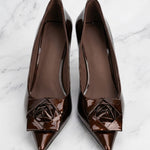 A pair of elegant dark chocolate brown patent leather pumps featuring a pointed toe and a sophisticated 3D rose floral decoration on the front, perfect for a quiet luxury office or evening look.